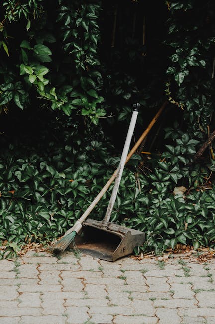 A black wheeled garden waste bin positioned on a grassy lawn, overflowing with assorted garden debris including dried grass, leaves, and twigs. The background features lush green bushes and trees, wit