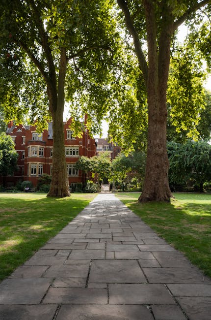 A paved stone pathway runs through a lush, well-maintained garden in Ravenscourt Park, Hammersmith, surrounded by vibrant green grass and tall trees with dense leafy canopies providing shade. In the b